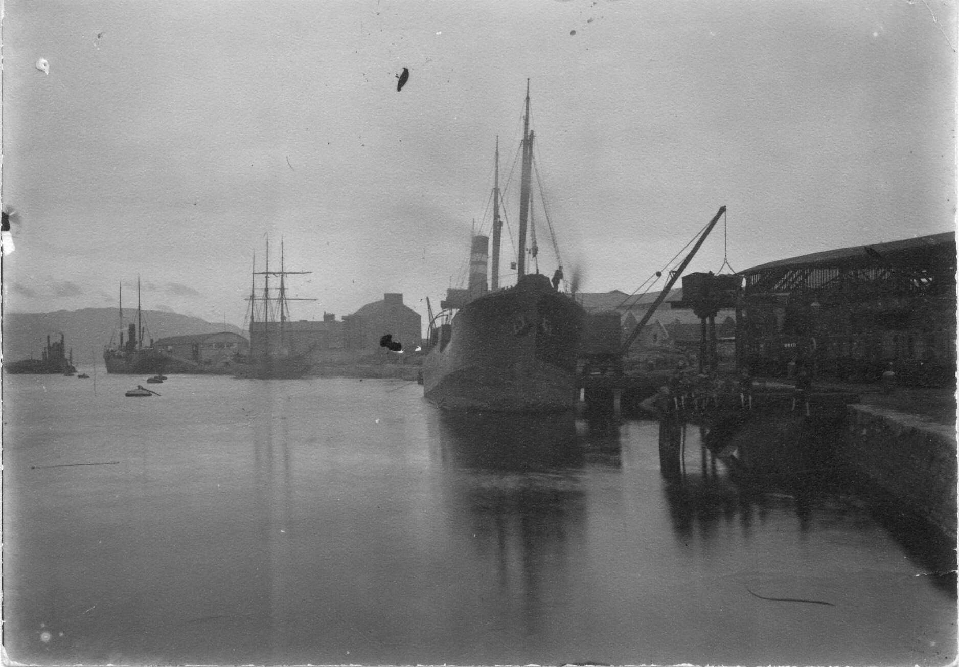 Dundalk harbour scene. Charlie McCarthy: showing from left-right: Dundalk Harbour Commissioners steam bucket dredger “Faugh-a- Ballagh” with hoppers alongside; steam passenger/livestock ship (probably the ss “Dundalk”); steampacket office and Jennings large store; a three mast sail vessel moored at Williamsons quay; a steam collier at Connicks quay with a steam crane discharging coal onto the overhead gantry and screened into railwagons underneath; and a group of children sitting on Browns quay. These quays, actually wooden jettys, were built over in the early 1930’s.